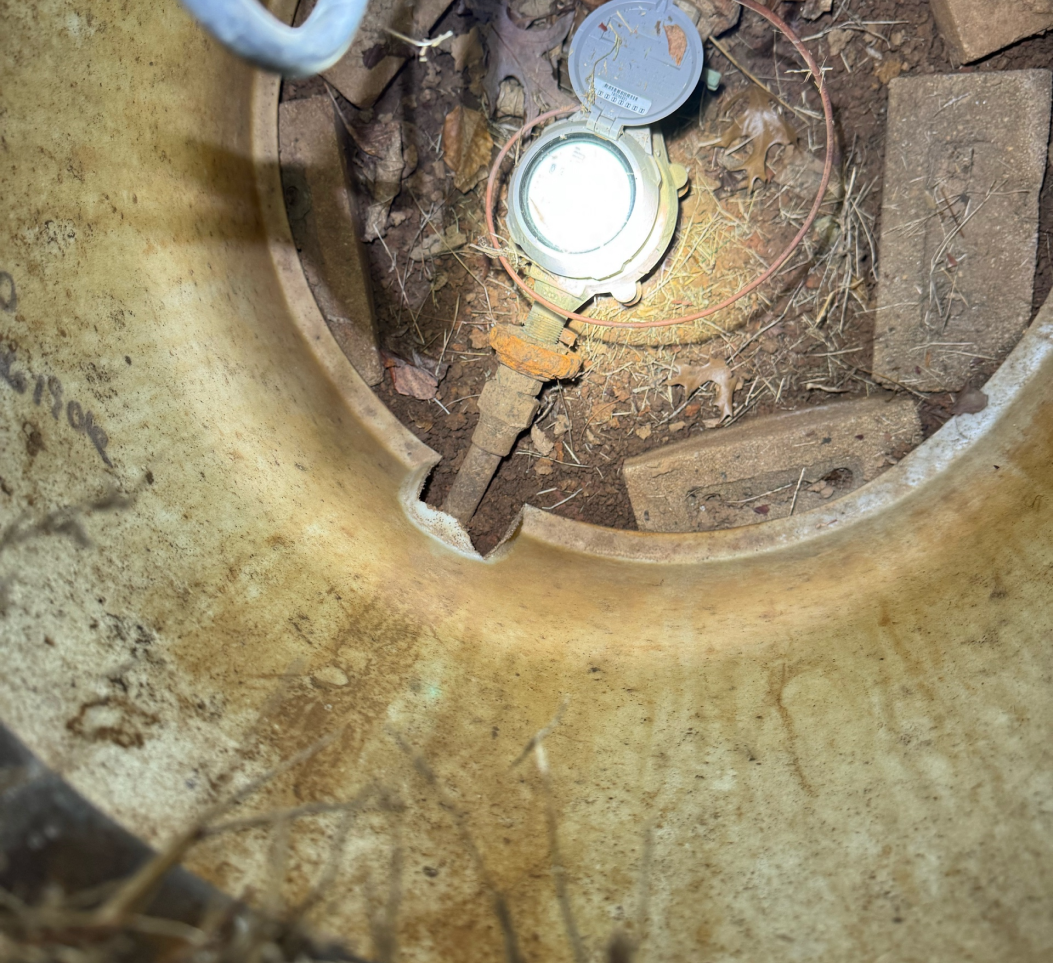 A close-up view of an exposed water meter with a glowing face, set in dirt and surrounded by leaves.