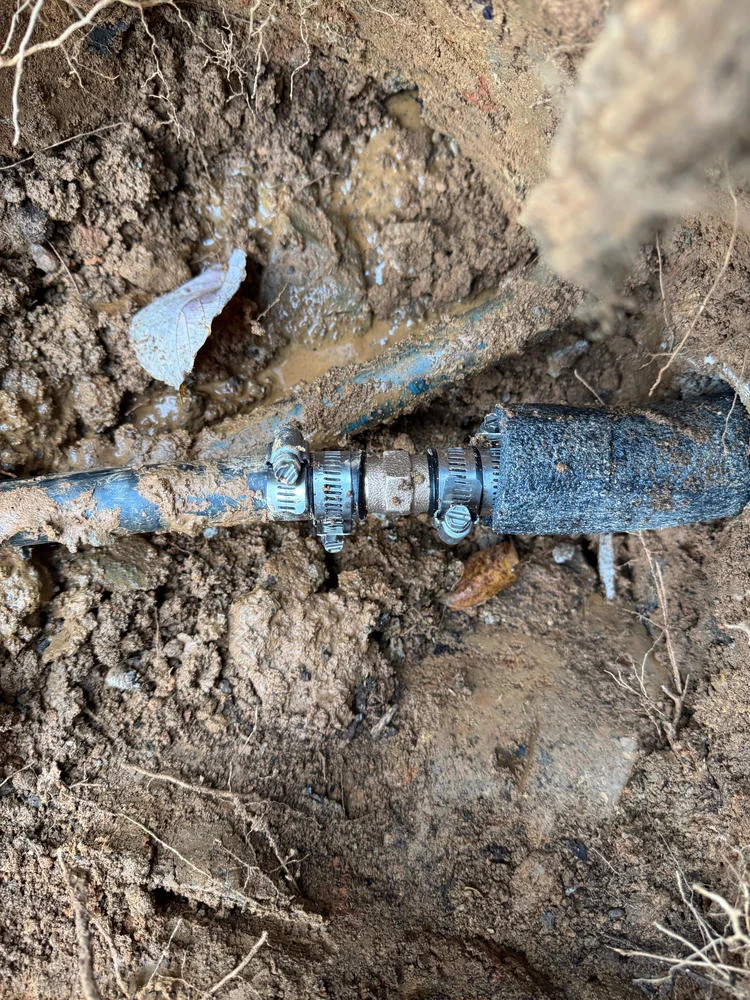 A black pipe with three metal clamps connecting two sections, surrounded by muddy brown soil and some roots.